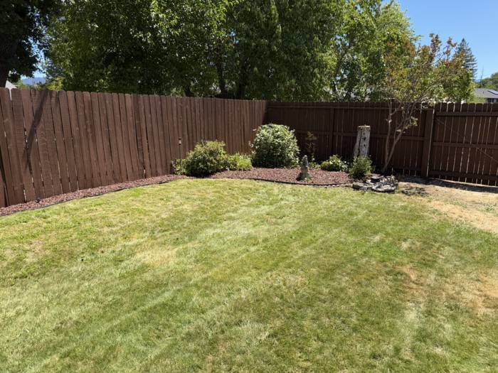 Backyard enclosed by a stained brown wood privacy fence, with a curved red lava rock planting bed and freshly mowed lawn in Grants Pass, Oregon.