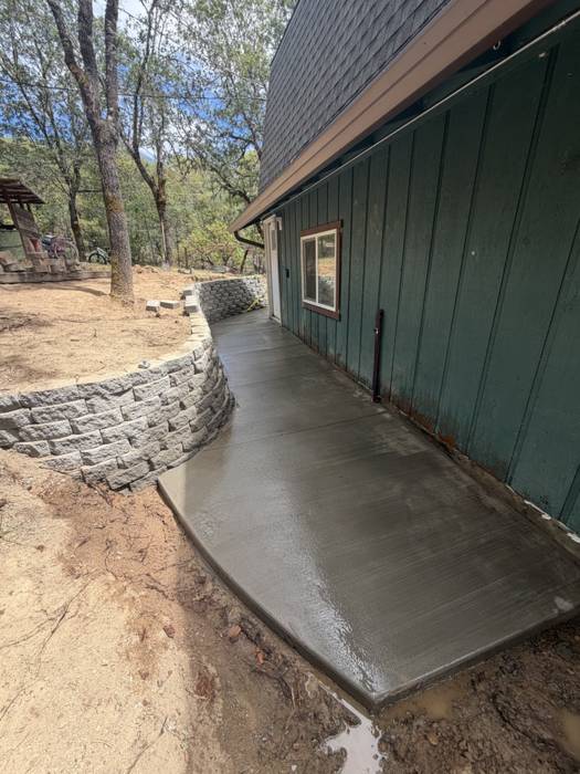 Newly poured broom-finish concrete walkway running along a green house, backed by a curved stacked-block retaining wall on a wooded Southern Oregon lot.