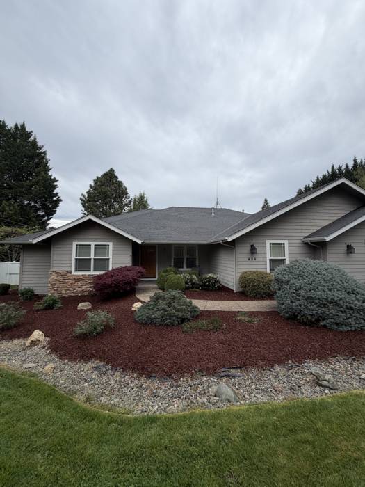 Finished front yard at a Grants Pass, Oregon home with fresh red lava rock beds, shaped evergreen and Japanese maple shrubs, a river rock dry border, and a cleanly edged lawn.