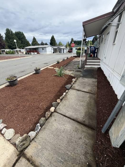 Bark mulch planting bed edged with a hand-set river rock border running alongside a concrete walkway at a Grants Pass, Oregon home.